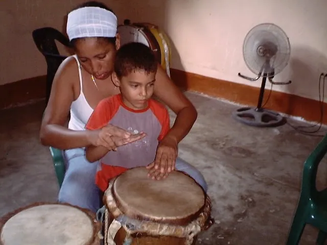 Un joven guía las manos de un niño sobre el cuero de la tambora en el taller de música tradicional.